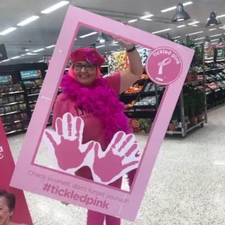 An ASDA colleague dressed in pink clothing, with a pink feather boa and beret holding a tickled pink instagram frame in an ADSA store | CoppaFeel!
