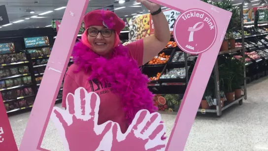 An ASDA colleague dressed in pink clothing, with a pink feather boa and beret holding a tickled pink instagram frame in an ADSA store
