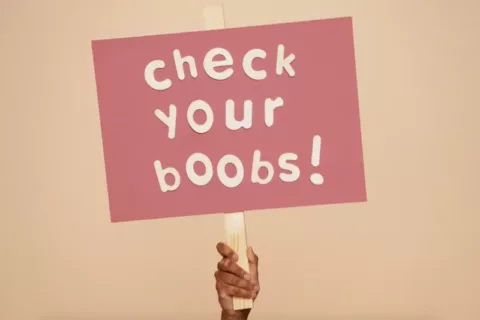 a pink sign that reads 'check your boobs!' being held up by a hand