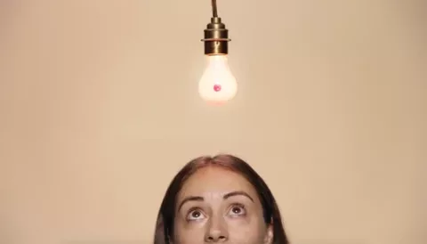 A woman is looking up in thought to a lightbulb that is lite and shaped like a boob