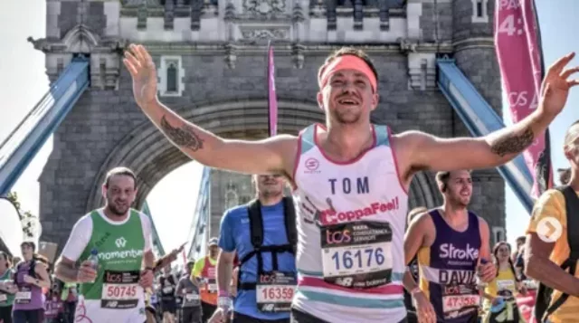 A group of runners crossing over tower bridge during a London marathon. The man at the front called tom is wearing a CoppaFeel! running vest and is smiling with his hands in the air