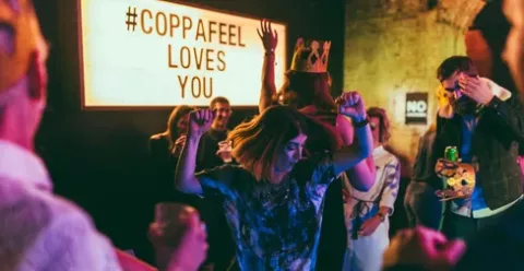 A group of people dancing in an evening venue in front of a light box sign that reads 'CoppaFeel! loves you'