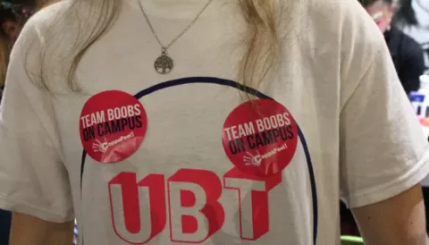 A close up of a person wearing a white t-shirt with 'UBT' written on it and two 'team boobs on campus' stickers on their chest