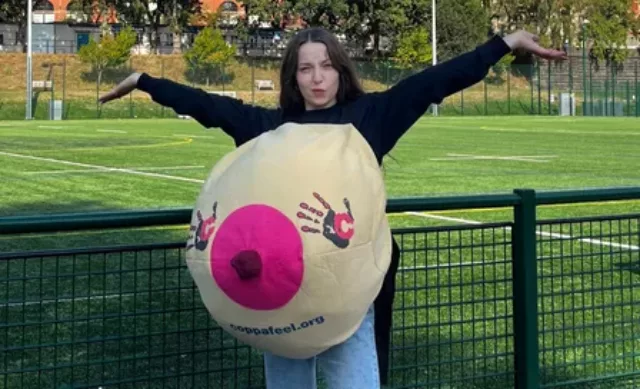 A woman from one of our university boob teams picture outside wearing a boob suit holding her hands in the air