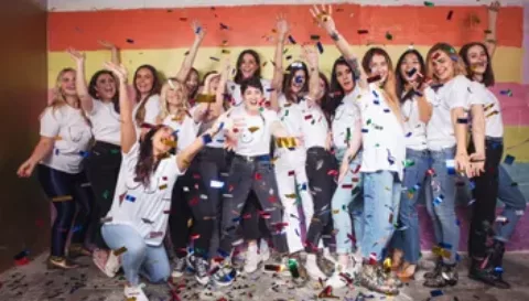 A group all wearing boob t-shirts celebrating by throwing multi-coloured confetti together