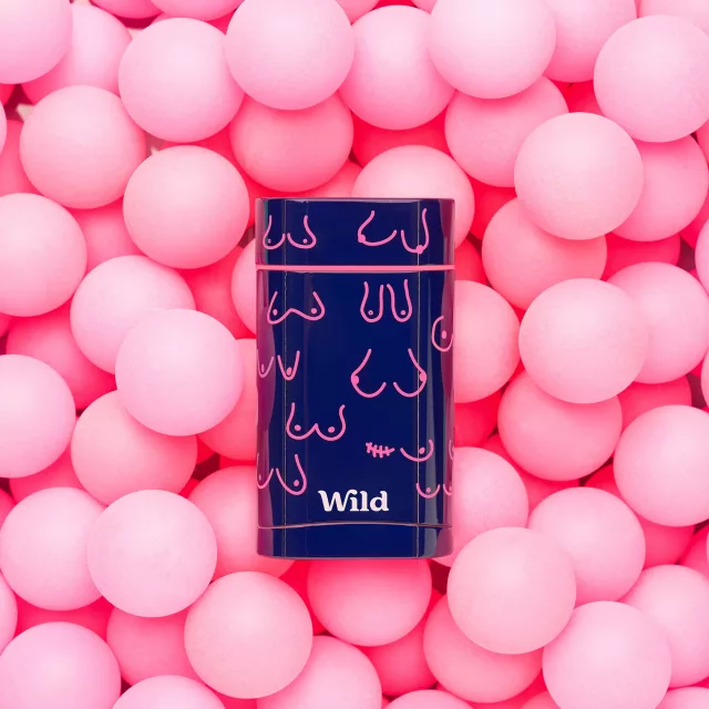 A navy reusable deodorant case with different chest, boob and pec illustrations in bright pink with the Wild Deodorant logo at the bottom in white. The case is sitting on top of a bright pink ball pit