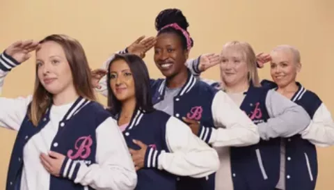 Five CoppaFeel! Boobettes standing in a line all saluting and holding their chests. The Boobettes are all wearing varsity style jackets with Boobette branding in CoppaFeel! navy and pink | CoppaFeel!
