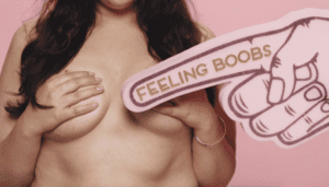 A close up of a woman clutching her chest with a pink foam finger that reads 'feeling boobs' pointing towards her chest | CoppaFeel!