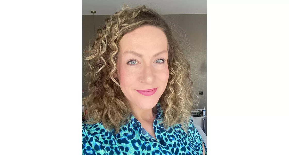 A white woman with brown curly hair smiling to camera. She is wearing a blue leopard printed top.