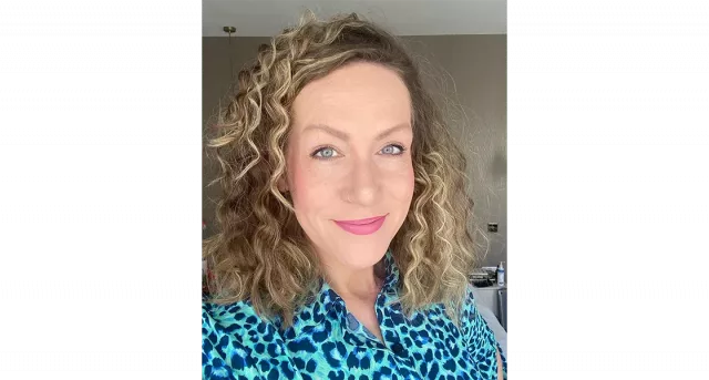 A white woman with brown curly hair smiling to camera. She is wearing a blue leopard printed top.