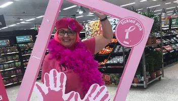 An ASDA colleague dressed in pink clothing, with a pink feather boa and beret holding a tickled pink instagram frame in an ADSA store