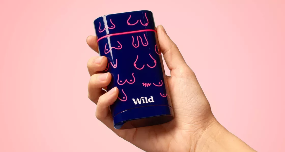 A navy reusable deodorant case with different chest, boob and pec illustrations in bright pink with the Wild Deodorant logo at the bottom in white. The case is being held up by a hand against a baby pink background