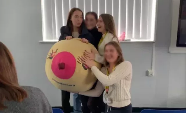 A group of young girls all stood around a boob suit hugging it and laughing