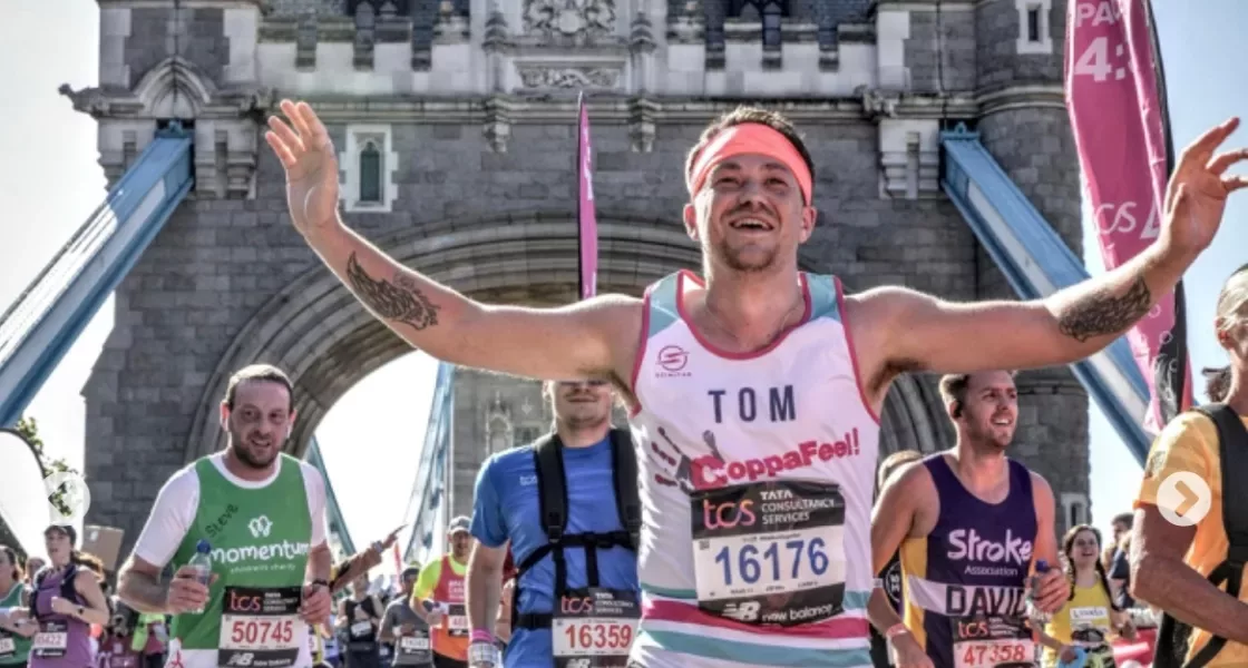 A group of runners crossing over tower bridge during a London marathon. The man at the front called tom is wearing a CoppaFeel! running vest and is smiling with his hands in the air