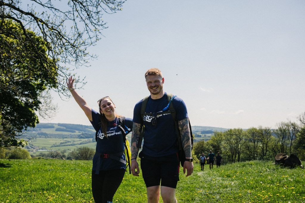 A man and woman walking during a CoppaTrek! challenge in field with clear blue skies. Both are wearing a navy blue CoppaTrek! t-shirt, both smiling and the woman is waving to camera