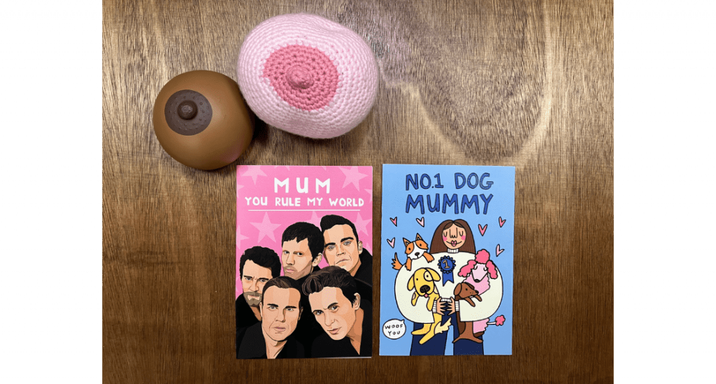 Two mothers day cards sit on top of a table. one card has an illustration of Take That on with the test 'Mum you rule my world' and the second card has an illustration of a woman holding lots of dogs, with the text 'NO1 Dog Mummy'. 