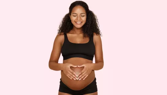A black woman seen in her underwear with her baby bump on show, she has her hands arranged in a heart over her bump